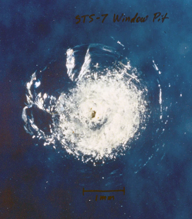 Archival NASA photograph shows an impact crater and cracking in a Space Shuttle window after the STS-7 mission